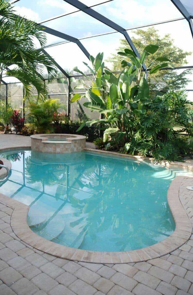 Indoor swimming pool features a kidney-shaped design with an integrated hot tub, surrounded by a paver stone deck and lush tropical greenery.