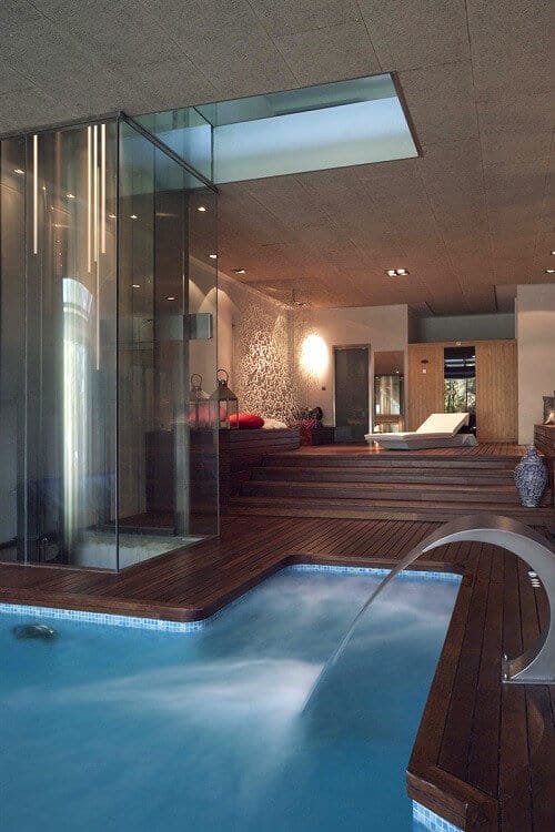 Rectangular indoor pool (10x5m) with smooth water, dark hardwood decking, glass walls for light, and modern lounge area, creating a spa-like vibe.