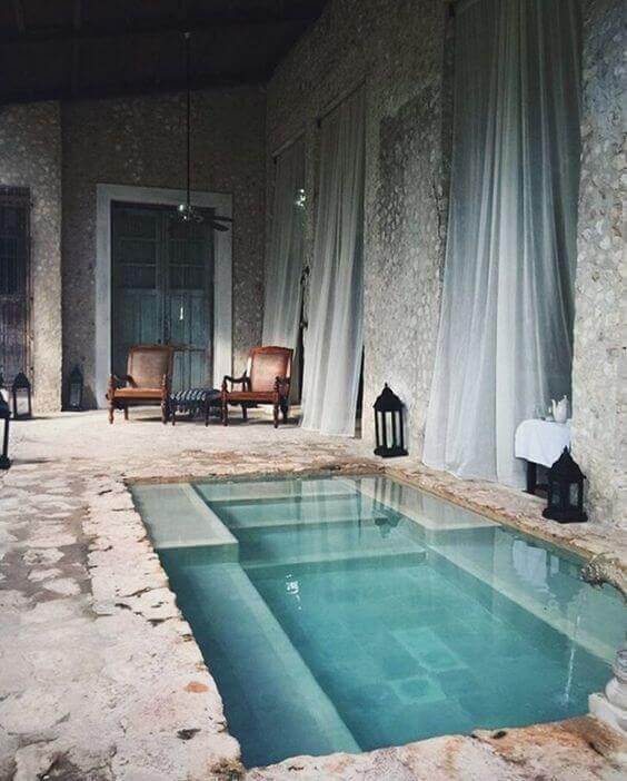 Indoor pool with a sunken design, surrounded by natural stone flooring. Features sheer curtains, wooden chairs, and high exposed beams.