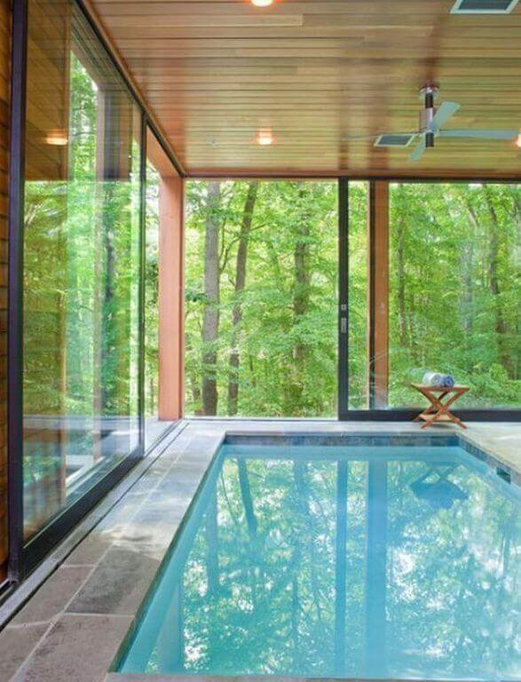 Sleek modern indoor swimming pool with floor-to-ceiling glass walls, light blue tiles, wood ceiling, and natural stone flooring, overlooking lush greenery.