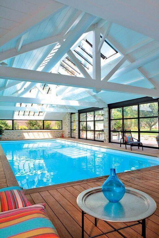 Indoor pool area with a polished light blue swimming pool, wooden deck, large glass walls, and cozy seating, blending indoor and outdoor spaces.