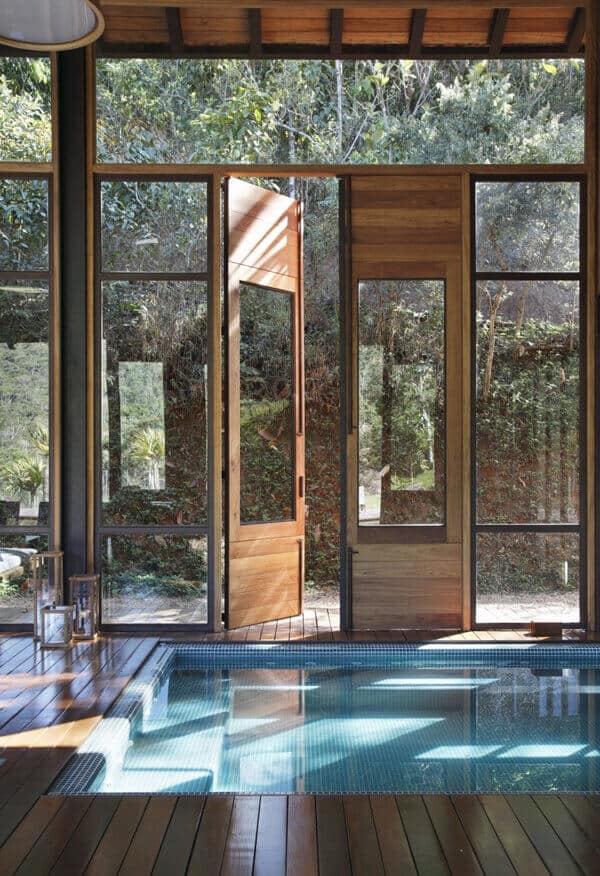 Sleek indoor pool measuring 12x6 feet, surrounded by dark wooden decking. Large glass doors open to lush greenery, enhancing the modern design.