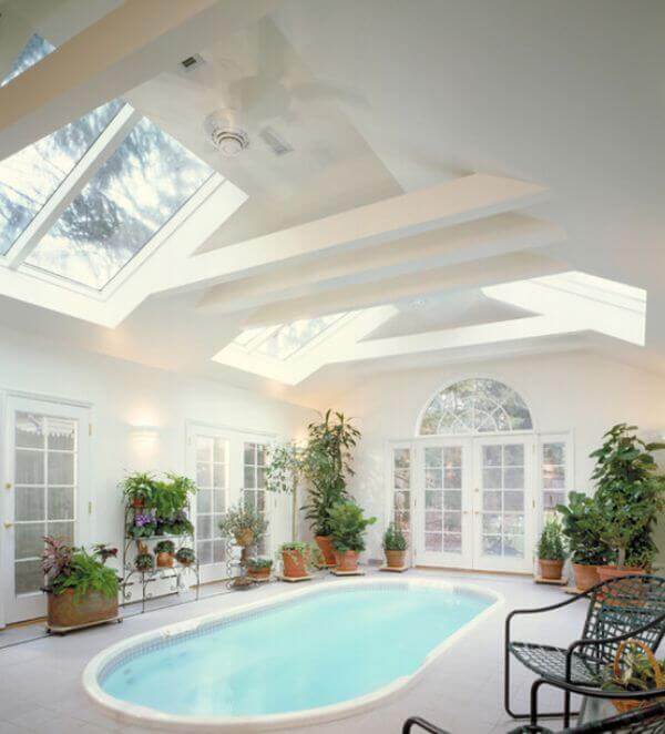 Indoor pool in a sunlit room with skylights, surrounded by a tiled deck and greenery in terracotta pots, blending relaxation and elegance.