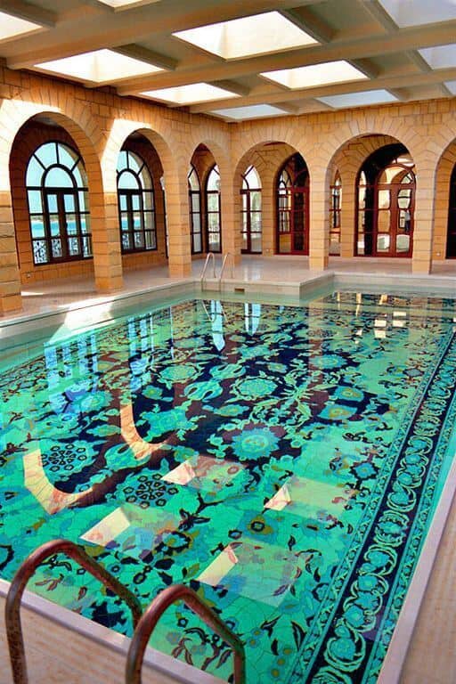 Indoor pool featuring intricate mosaic tile work in vibrant greens and blues, arched stone columns, and large windows for natural light.