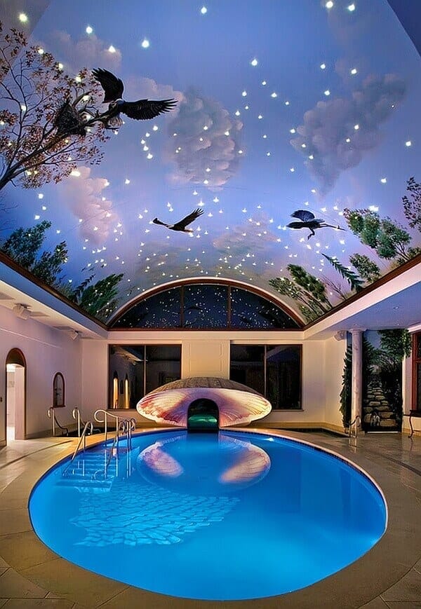Sprawling oval indoor pool with shimmering blue water, surrounded by grey stone tiles, featuring a shell-shaped spa and starry mural above.