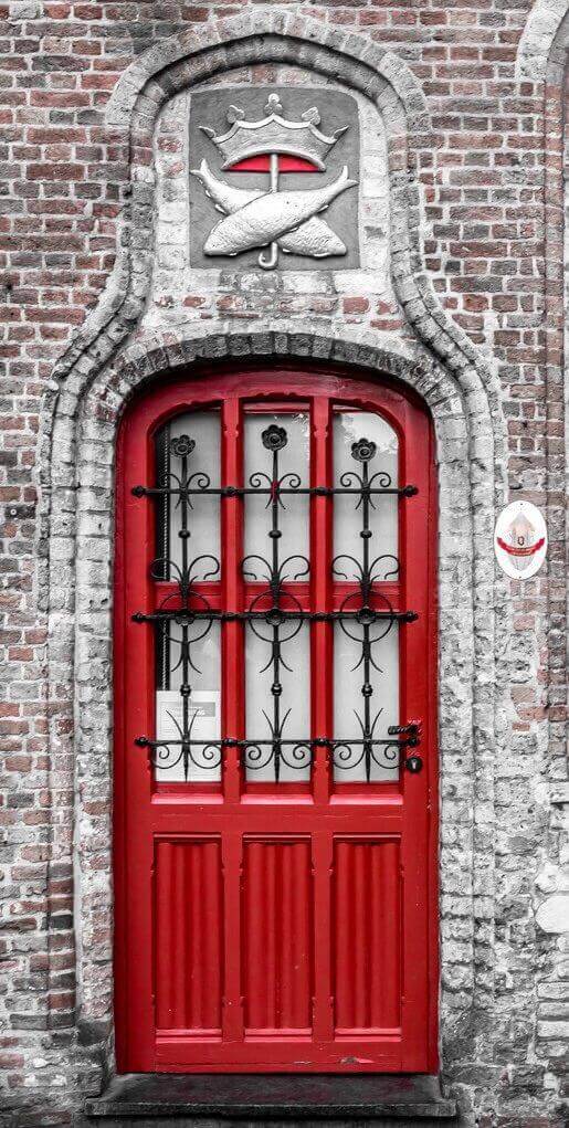 Bold red front door with ornate wrought iron details, double-panelled ribbed texture, set in a textured brick wall with a decorative crest.