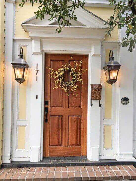 Front door design with rich wooden finish, six panels, elegant wreath, classic lanterns, light yellow siding, and welcoming brick steps.