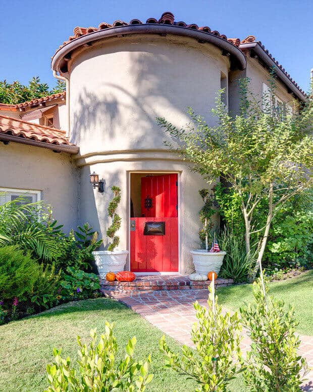 Vibrant red front door on a turreted home, surrounded by greenery, seasonal pumpkins, and a brick pathway, showcasing modern exterior door design.