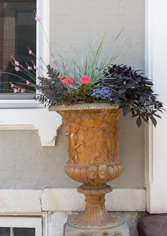 Ornamental planter, 3 feet tall, with intricate carvings. Filled with vibrant plants: tall grasses, pink and purple flowers, and burgundy foliage.