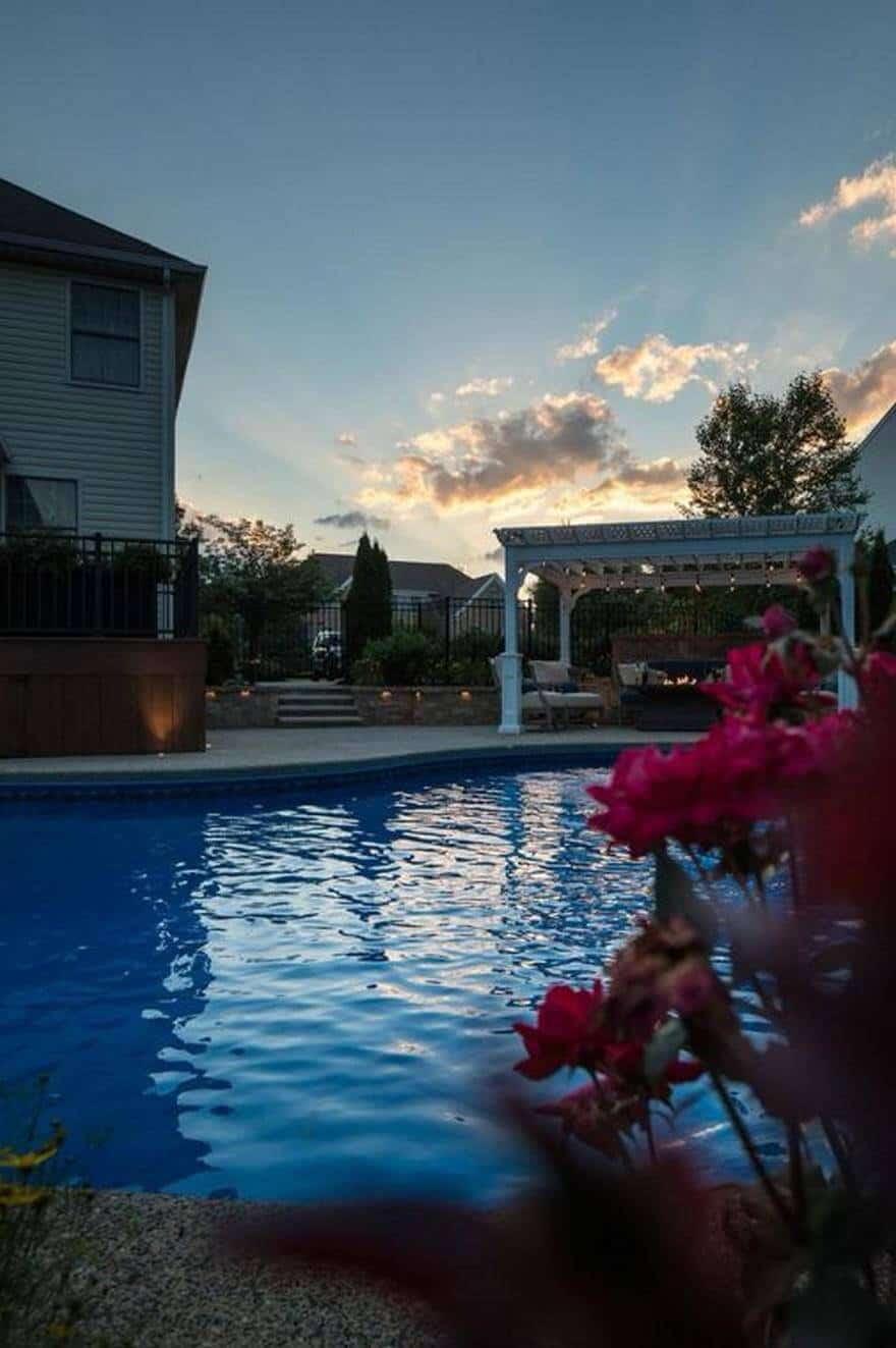 25 Pool Pergola Ideas for a Relaxing Oasis 2 Backyard pool features a rectangular design with blue tiles, surrounded by a wooden deck. A white pergola with soft lighting offers shaded seating, complemented by vibrant pink flowers along the edge.
