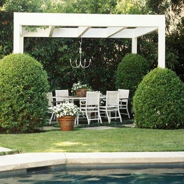 25 Pool Pergola Ideas for a Relaxing Oasis 13 Outdoor dining area with a white wooden pergola over a rectangular table, surrounded by lush greenery, next to a swimming pool.