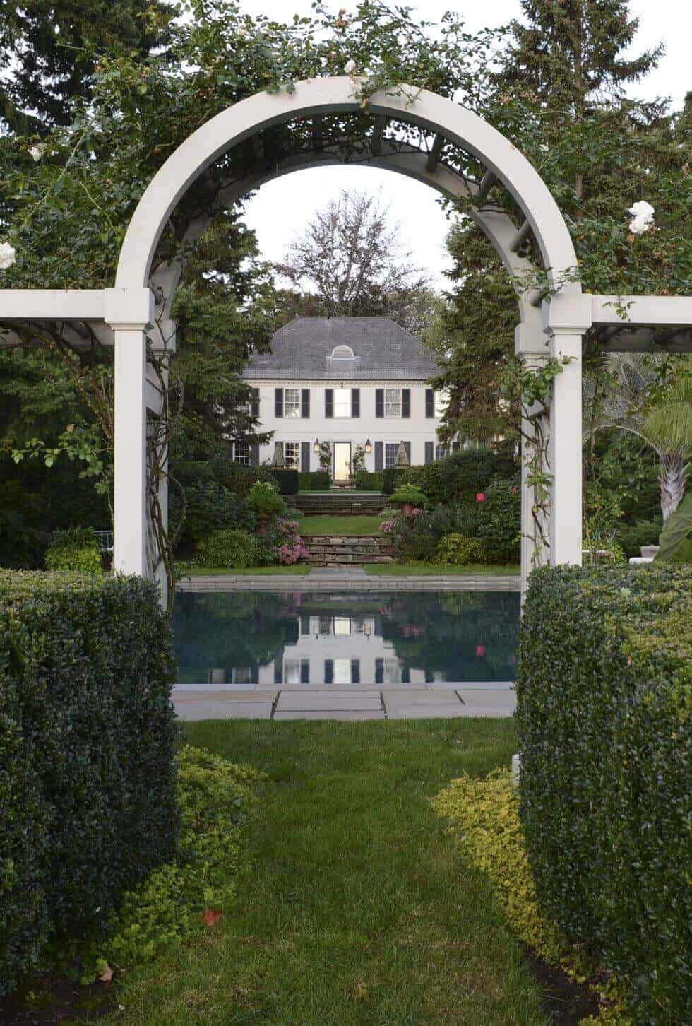 25 Pool Pergola Ideas for a Relaxing Oasis 15 Landscaped garden with a central archway leading to a serene pool, surrounded by hedges and flowers, complemented by a classic colonial house.