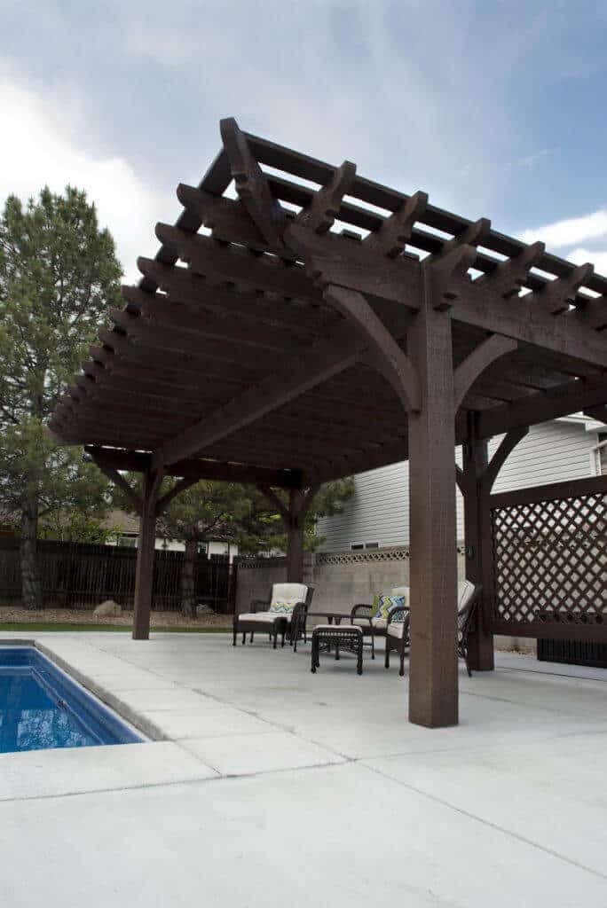 25 Pool Pergola Ideas for a Relaxing Oasis 18 Sturdy wooden pergola over a backyard pool, featuring dark wood contrasting with a light paved deck, providing shade for seating.