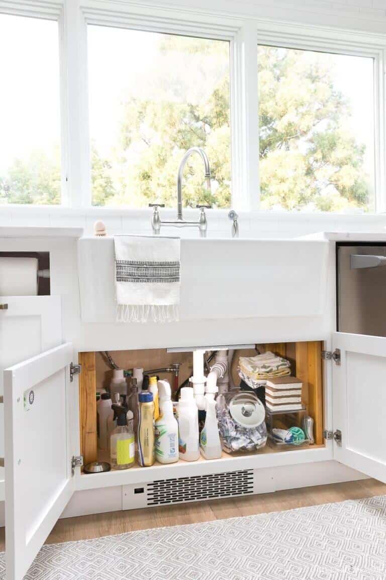 Well-organized under-sink storage with white cabinetry, showcasing bottles on a wooden shelf, blending functionality and modern farmhouse style.