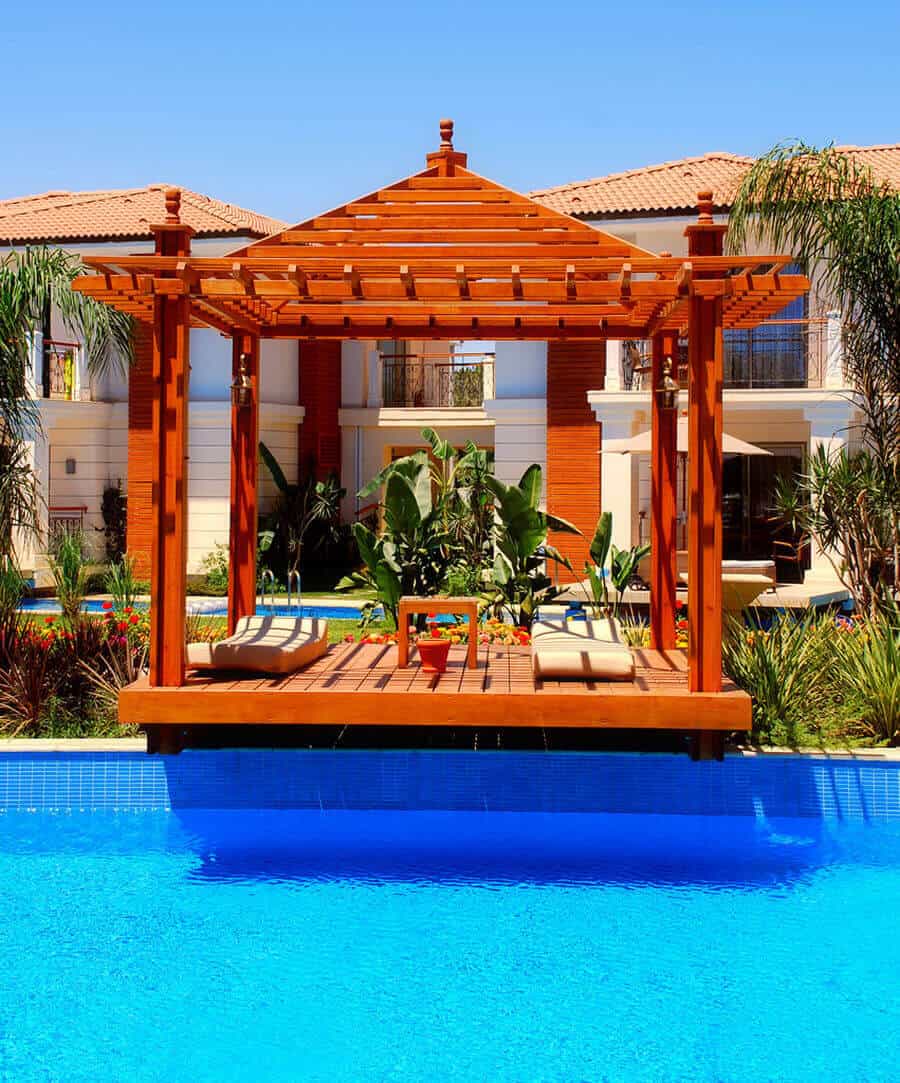 25 Pool Pergola Ideas for a Relaxing Oasis 21 Wooden pergola with mahogany finish over a rectangular pool. Weather-resistant timber deck and tropical plants enhance the outdoor space.