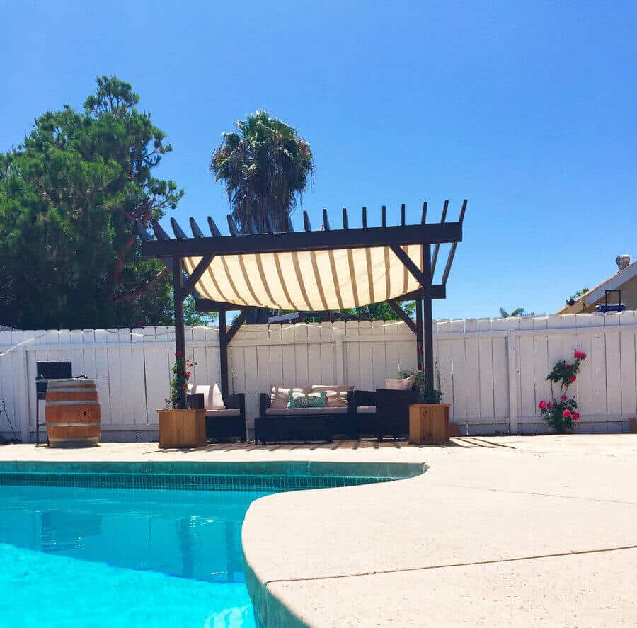 25 Pool Pergola Ideas for a Relaxing Oasis 23 Wooden outdoor pergola by the pool, 10x12 feet, with slatted roof for shade. Cushioned seating and vibrant landscaping create an inviting retreat.