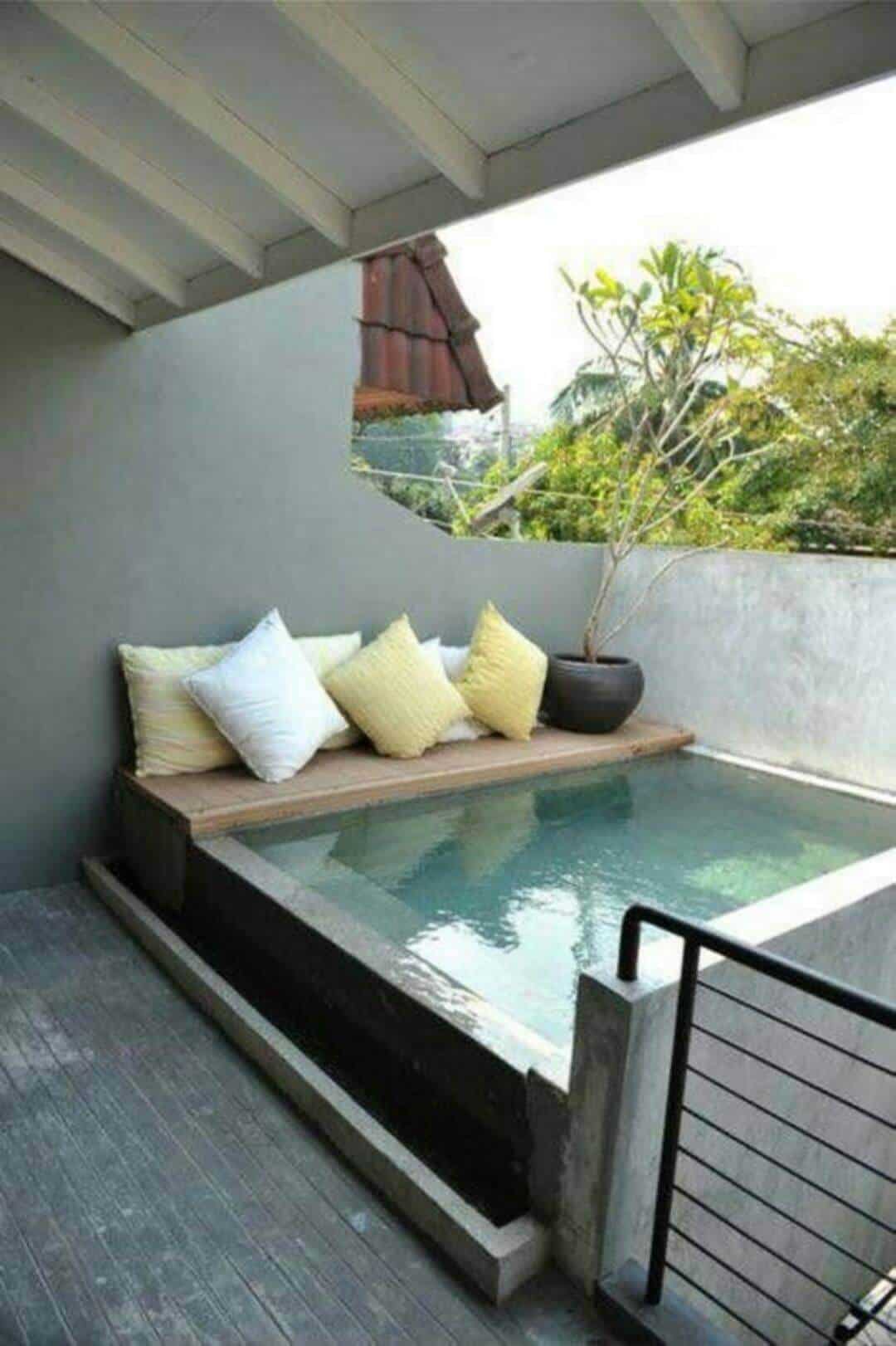 25 Pool Pergola Ideas for a Relaxing Oasis 24 Wooden platform adjacent to a small plunge pool, adorned with soft pillows, surrounded by light gray walls, offering a private, shaded outdoor lounging area.