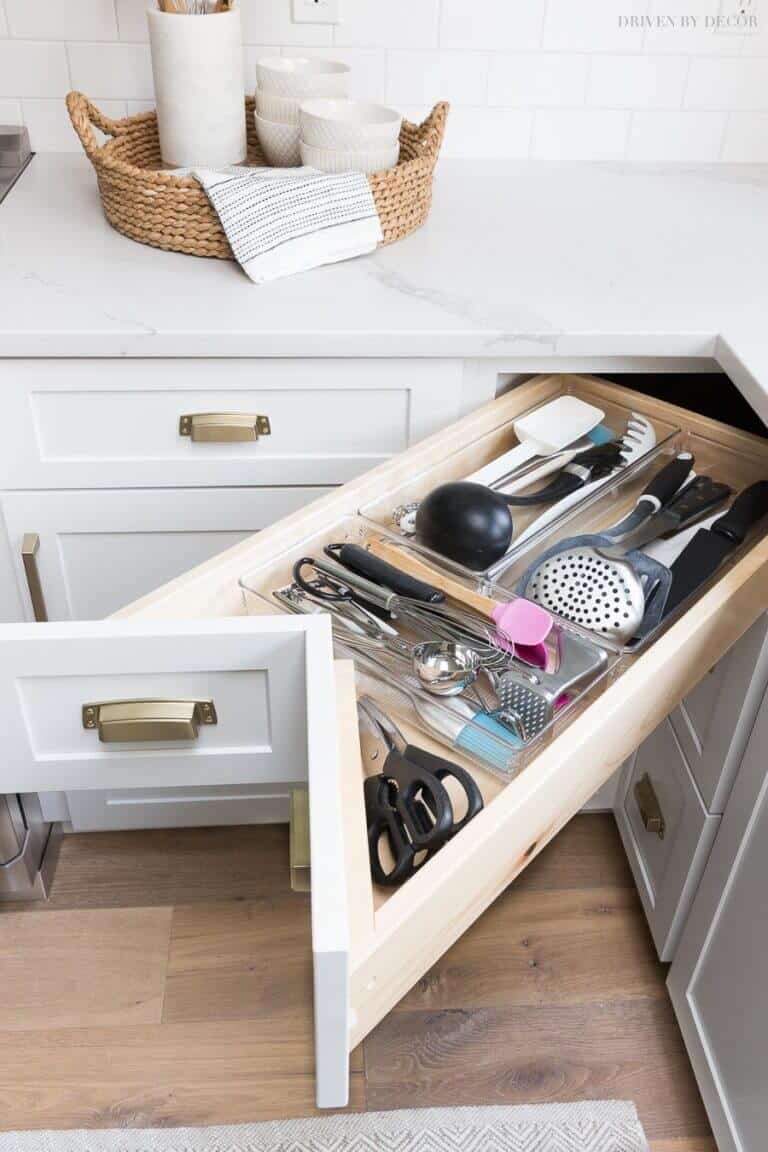 Neatly organized kitchen drawer with utensils like whisks and spatulas in clear organizers. Elegant white cabinetry and brass handles.