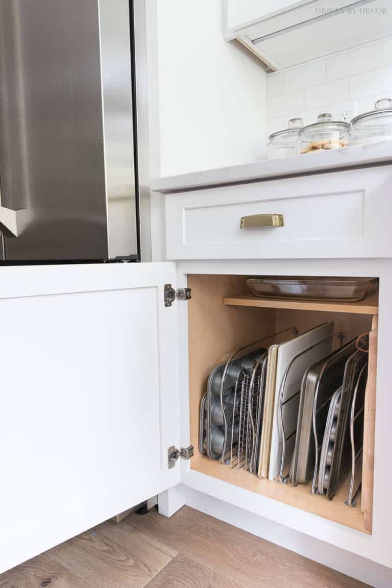 Neatly organized kitchen cabinet with white shaker-style doors and gold handles, displaying stacked baking sheets and pans for optimal storage.