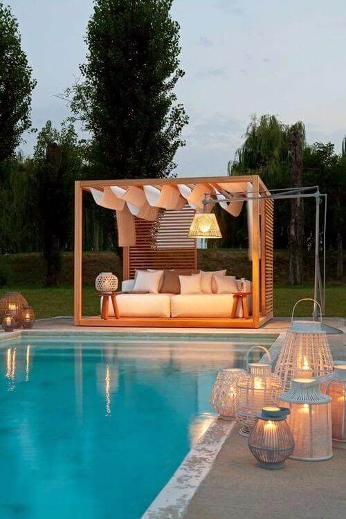 25 Pool Pergola Ideas for a Relaxing Oasis 6 Outdoor lounge with a wooden frame by a pool, featuring a cushioned white sofa with decorative pillows, ambient lighting, and lush greenery.