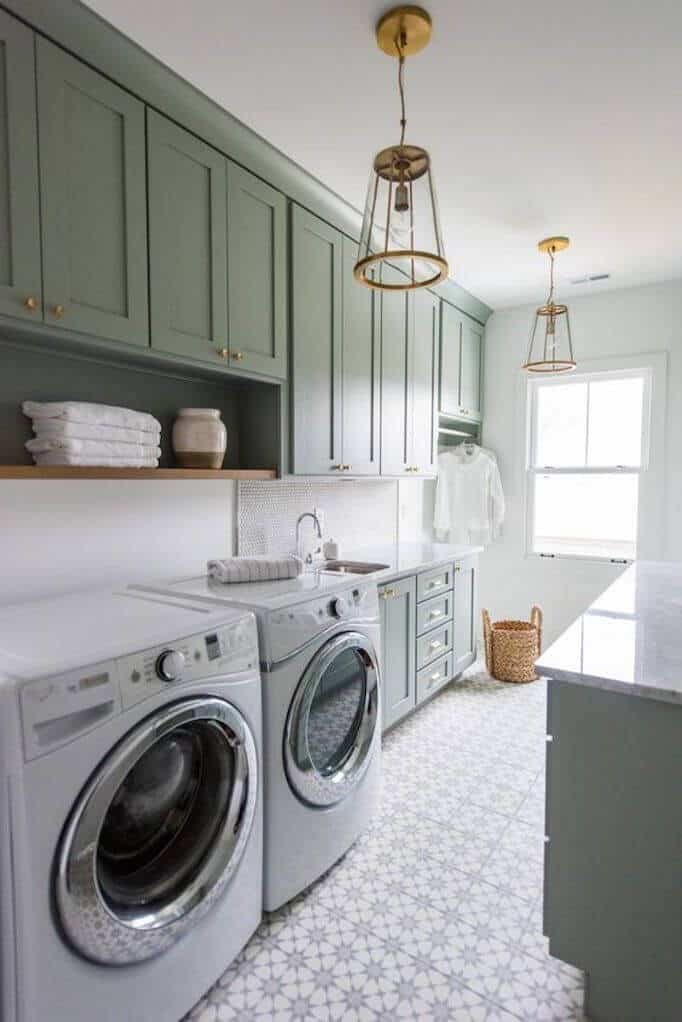 Sage green cabinetry with gold hardware complements wall-mounted shelves in a modern laundry room, illuminated by pendant lights.