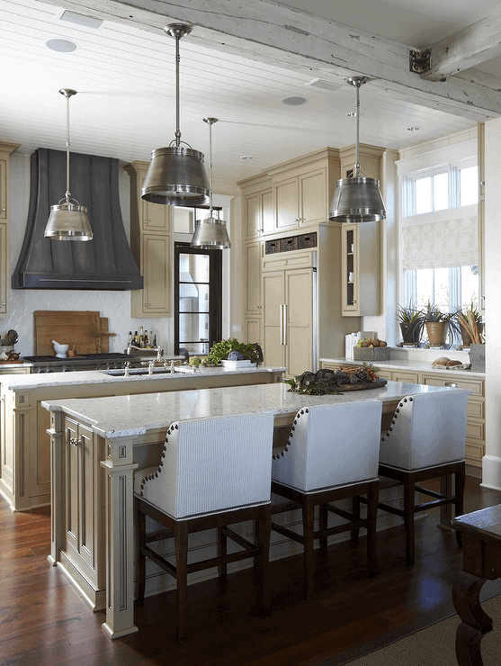 Modern kitchen with two marble islands, cream cabinetry, dark wood flooring, and minimalist white chairs. Brightened by large windows and pendant lights.