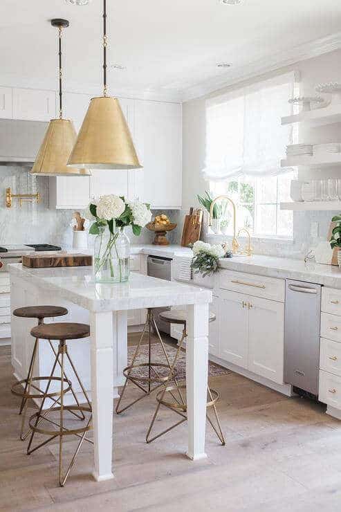 Modern kitchen with a white marbled island, three brass-accented stools, and pendant lighting. Sleek cabinetry and warm wooden flooring enhance functionality.
