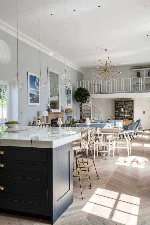 Modern kitchen with a large white marble island, navy blue base, mixed seating, herringbone wooden flooring, and ample natural light.