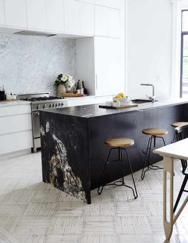 Modern kitchen with a black and white color scheme. Features a large textured black stone island, minimalist wooden stools, and glossy white cabinetry.