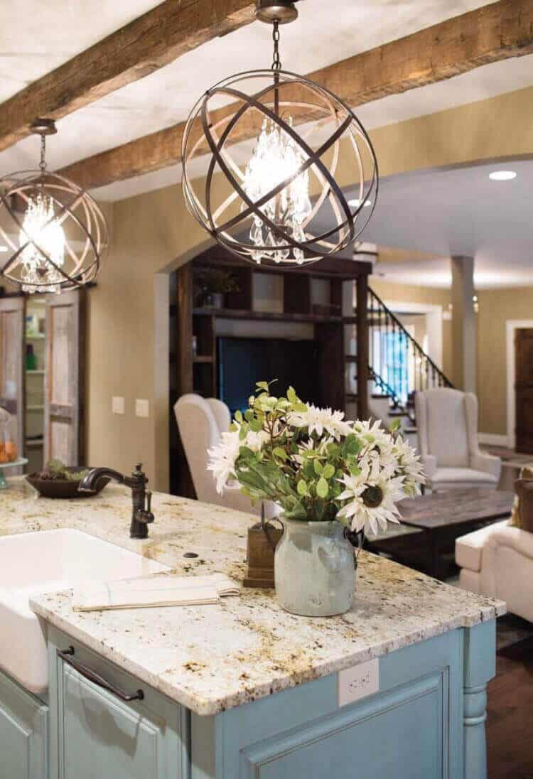 Central kitchen island with a granite countertop in soft blue, pendant lights above, rustic beams, and integrated stainless-steel sink.