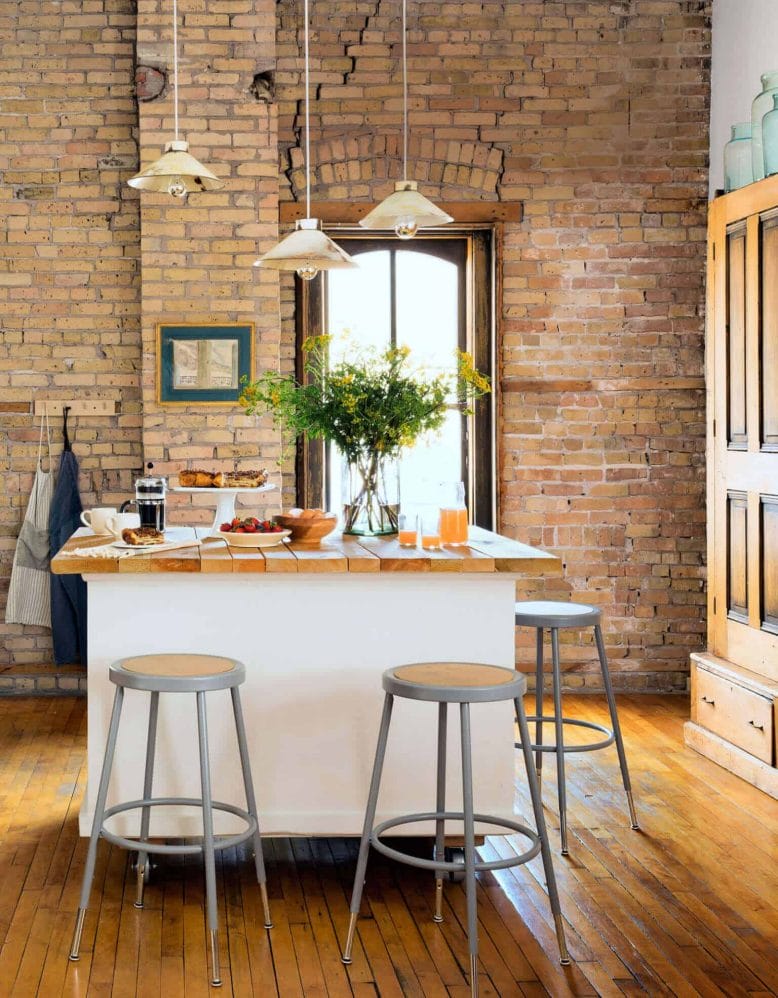 Central kitchen island with wooden countertop, white cabinetry, three pendant lights, and exposed brick walls, surrounded by metal bar stools.