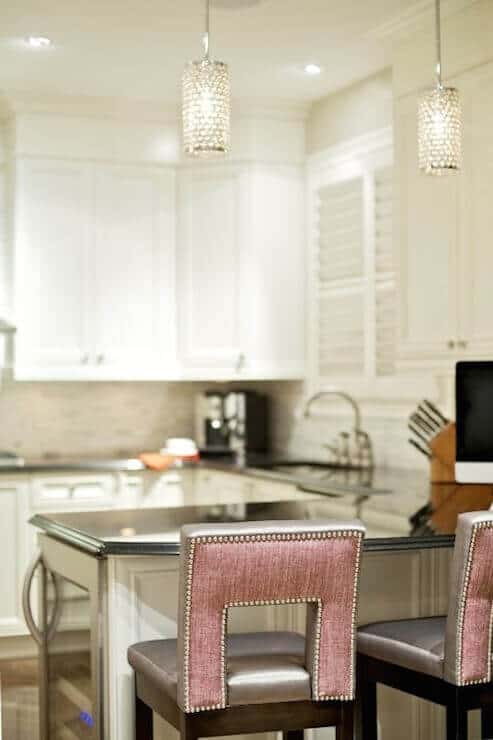 Modern kitchen with sleek white cabinetry and a 6-foot dark island countertop. Features recessed and pendant lighting, a built-in sink, and pink upholstered bar stools. Natural light adds warmth to the functional layout.
