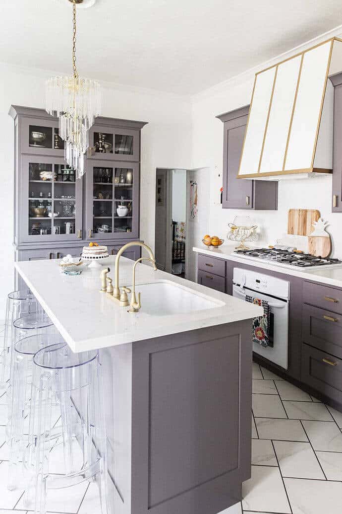 Modern kitchen featuring an 8-foot white marble island with transparent acrylic bar stools, muted gray cabinetry, and a crystal chandelier.